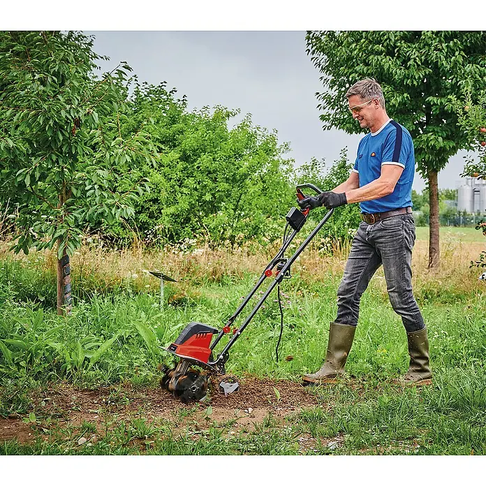 Einhell Azada eléctrica GC-RT 1545 M - 1.500 W, Anchura de trabajo: 45 cm, Profundidad de trabajo: 12 cm - 22 cm - Un hombre está usando una motoazada en el jardín para aflojar la tierra. La motoazada es roja y negra y parece fácil de usar.