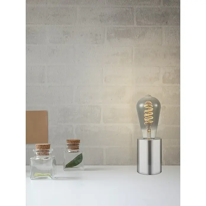 Home Sweet Home Lampenfuß Dry - 10 W, E27, Stahl, Höhe: 10 cm - Nahaufnahme einer silbernen Lampe mit Edison-Glühbirne auf einem weißen Tisch, neben kleinen Glasflaschen und einer beigen Karte.