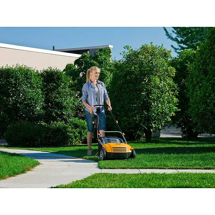 Stiga Elektro-Vertikutierer SV 213 E - 1.300 W, Arbeitsbreite: 32 cm - Eine blonde Frau mäht mit einem gelb-schwarzen Rasenlüfter den Rasen entlang eines Gehwegs, umgeben von üppigem Grün.