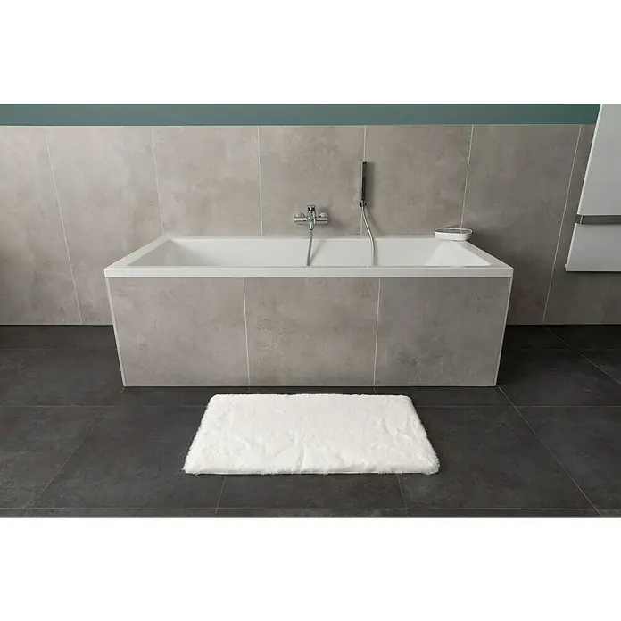 Badteppich Happy - 50 x 90 cm, Weiß, 100% Polyester - Badezimmer mit einer modernen Badewanne, umgeben von grauen Fliesen, und einer flauschigen, weißen Badematte auf dunklen Bodenfliesen.