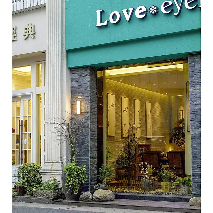 Starlux Laholm LED-Außenwandleuchte - 8 W, 7 x 5,3 x 30 cm, Dunkelgrau, IP54 - Außenansicht eines Geschäfts mit dem Namen "Love Eye", dekoriert mit Pflanzen und Steinelementen.