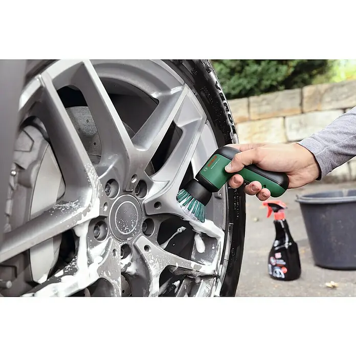 Bosch Akku-Reinigungsbürste UniversalBrush - 14 -tlg., 3,6 V, 2 Ah - Eine Person reinigt eine silberne Autofelge mit einer grünen Bosch Bürste. Reinigungsmittel sind auch im Bild zu sehen.
