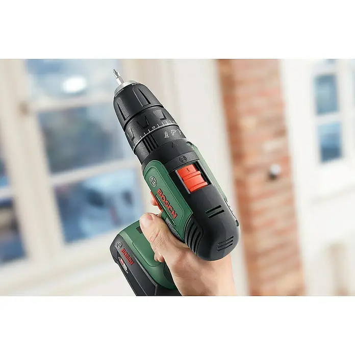 Bosch Power for All 18V Taladro atornillador percutor de batería UniversalImpact 18 - 18 V, 2 baterías, 1,5 Ah, 34 Nm - Primer plano de un taladro inalámbrico Bosch con una broca reemplazable, sostenido en una mano contra un fondo brillante.