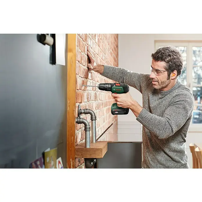 Bosch Power for All 18V Taladro atornillador percutor de batería UniversalImpact 18 - 18 V, 2 baterías, 1,5 Ah, 34 Nm - Un hombre perfora una pared de ladrillo con un taladro inalámbrico Bosch verde. Lleva gafas de seguridad y tapones para los oídos.