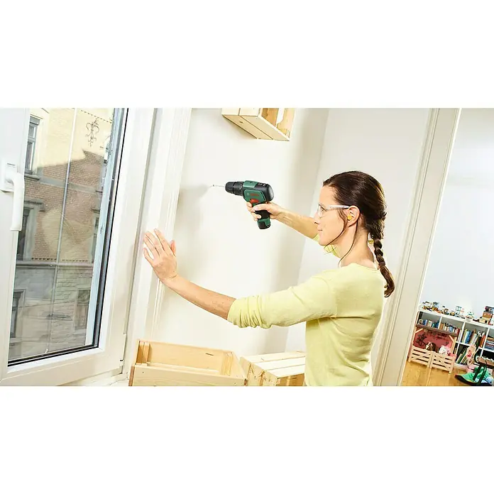 Bosch Power for All 12V Akku-Schlagbohrschrauber EasyImpact 12 - 12 V, 1 Akku, 2 Ah - Eine Frau mit Schutzbrille benutzt einen Bosch Akku-Bohrschrauber, um eine Holzkiste an einer weißen Wand zu befestigen.