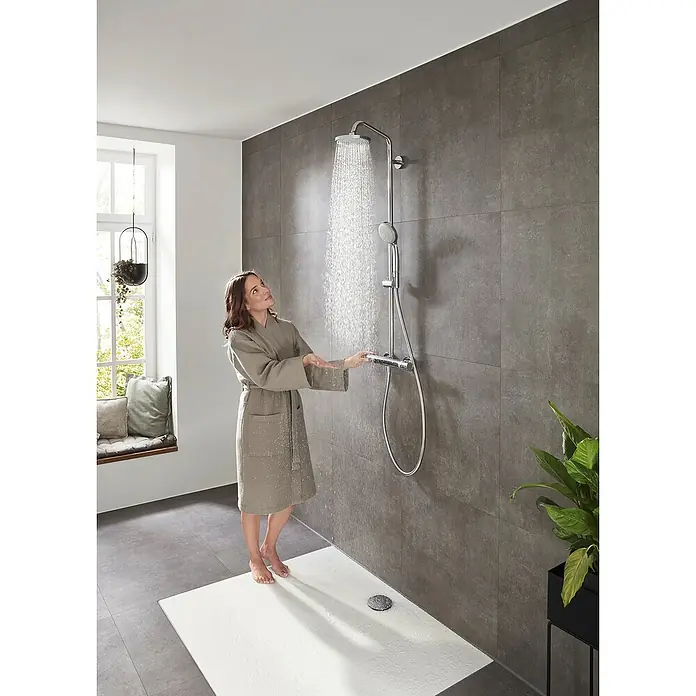 Hansgrohe Duschsystem Croma Showerpipe 160 - Mit Thermostatarmatur, Lochabstand: 93,2 cm, Anzahl Strahlarten: 4 Stk., Chrom, Höhe: 110,1 cm - Eine Frau im Bademantel testet eine moderne, verchromte Duscharmatur mit großem Duschkopf und Handbrause vor einer grauen Wand.