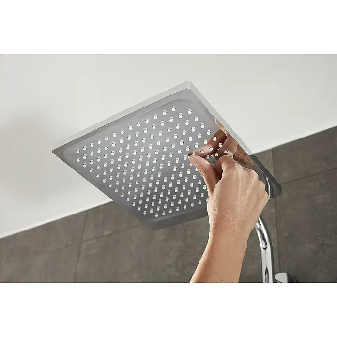 Hansgrohe Crometta Duschsystem E 240 - Mit Thermostatarmatur, Lochabstand: 103,4 cm, Anzahl Strahlarten: 2 Stk., Chrom - Nahaufnahme eines quadratischen Duschkopfes aus Edelstahl, der von einer Hand gereinigt wird, um Kalkablagerungen zu entfernen.
