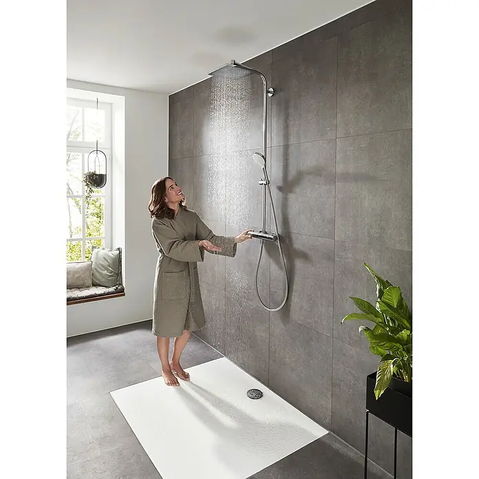 Hansgrohe Crometta Duschsystem E 240 - Mit Thermostatarmatur, Lochabstand: 103,4 cm, Anzahl Strahlarten: 2 Stk., Chrom - Eine Frau im Bademantel steht unter einer modernen Regendusche mit Handbrause und prüft die Wassertemperatur. Elegantes Baddesign mit grauen Fliesen.