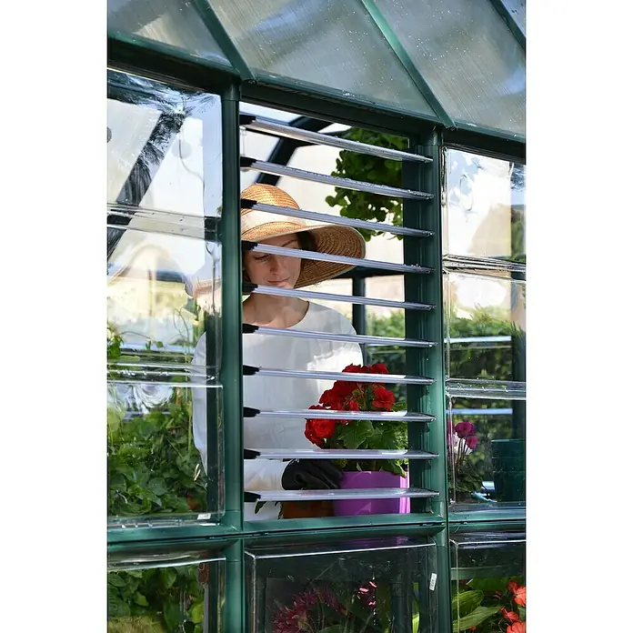 Palram – Canopia Ventana lateral para invernadero - Específico para: invernaderos Palram - Canopia - Una mujer en un invernadero con un sombrero, sosteniendo una planta con flores rojas en una maceta morada vista a través de las lamas.