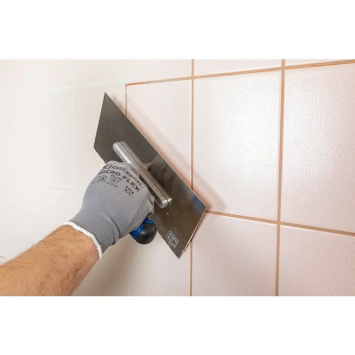 Knauf Rotband Flächenspachtel Plus - 4 kg - Nahaufnahme einer Person, die mit einer Glättkelle Fugenmörtel zwischen hellrosa Fliesen aufträgt, wobei der Fokus auf der Kelle und der Hand liegt.