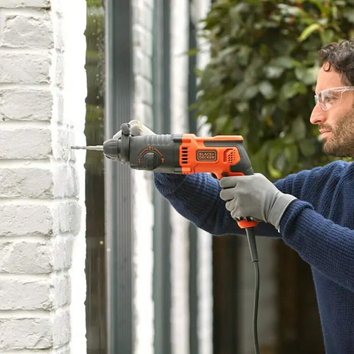 Black+Decker Martillo combinado BEHS01K-QS - 650 W, 1,4 J, Alojamiento SDS-Plus - Un hombre usa un taladro percutor Black+Decker para perforar una pared de ladrillo blanco. Lleva gafas de seguridad y guantes.