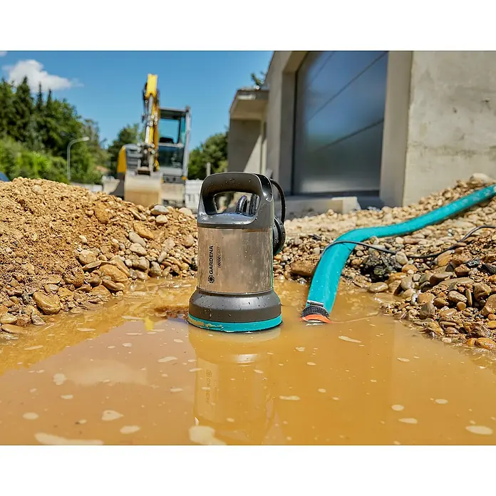 Gardena Bomba para aguas residuales 25000 - 1.100 W, Capacidad de bombeo máx.: 25.000 l/h - La bomba sumergible para aguas sucias GARDENA 18000 Dirt en acción, bombeando agua de obra, rodeada de piedras y barro.