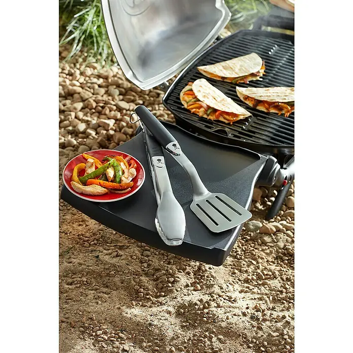Weber Utensilios para barbacoa Compacto - 2 pzs., Acero inoxidable - Una parrilla Weber portátil con quesadillas, pinzas, espátula y un plato de verduras a la parrilla, todo sobre un suelo pedregoso.