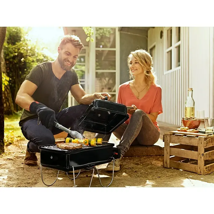 Weber Go-Anywhere Gasgrill - Grillfläche (B x T): 42 x 26 cm, 1,9 kW, Anzahl Brenner: 1 - Ein Paar grillt im Freien mit einem tragbaren Grill. Der Grill ist schwarz und hat einen Deckel, der geöffnet ist, um das Essen zu wenden.