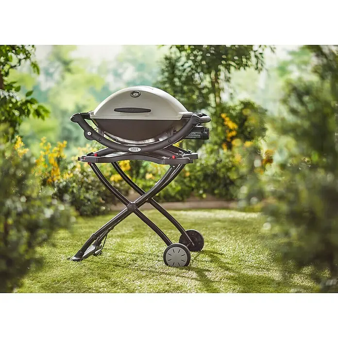 Weber Carro con ruedas para barbacoa - Específico para: Serie de barbacoa Q1000 / Q2000 - Una parrilla de gas Weber Q 1200 en un carrito con ruedas al aire libre, con un diseño gris y negro, rodeada de vegetación.
