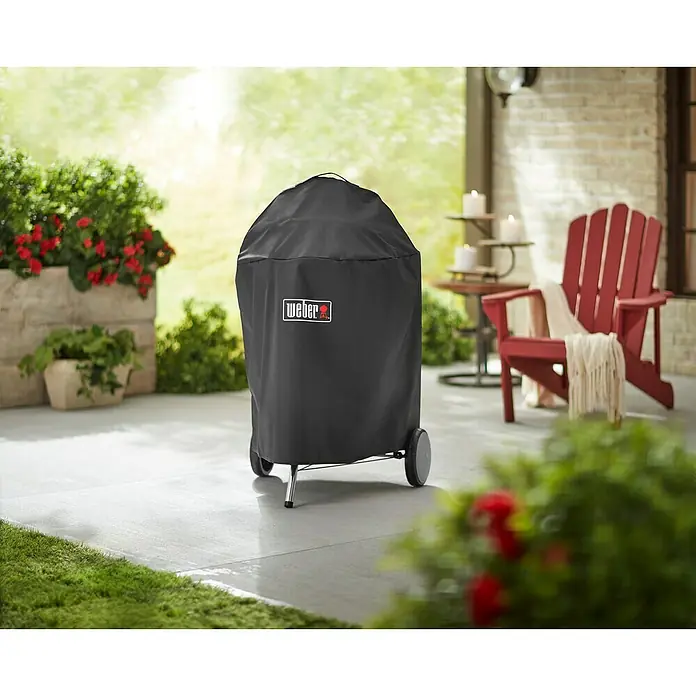 Weber Grill-Schutzhülle Premium - Passend für: Weber Holzkohlegrill mit Ø 57 cm - Ein Weber-Grill mit Schutzhülle auf einer Terrasse, umgeben von Pflanzen und einem roten Stuhl. Die Hülle schützt den Grill vor Witterungseinflüssen.
