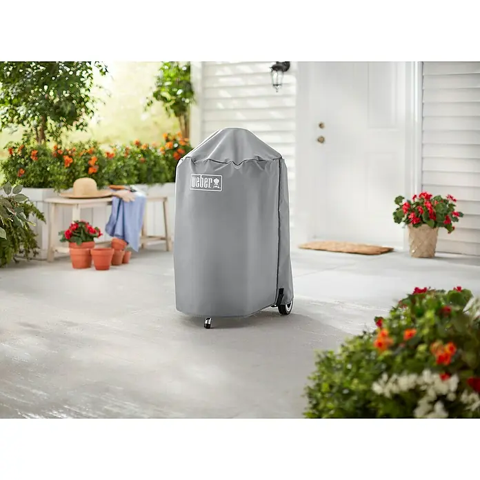 Weber Grill-Schutzhülle - Kunststoff, Passend für: Weber Kugelgrill Ø 47 cm - Das Bild zeigt eine graue Weber Grillabdeckung, die einen Grill schützt, auf einer Terrasse mit Pflanzen und Blumen im Hintergrund.