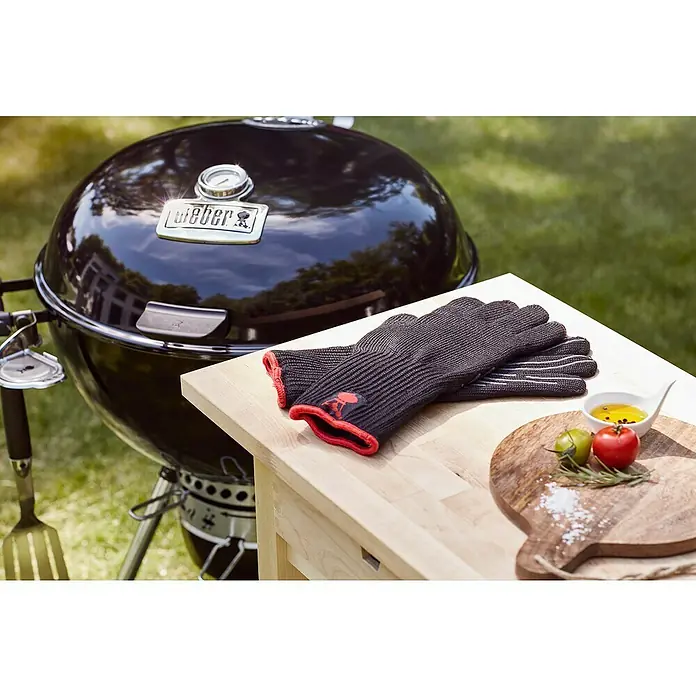 Weber Master-Touch GBS Kugelgrill Premium E-5770 - Durchmesser Grillfläche: 57 cm, Schwarz/Silber - Das Bild zeigt Grillhandschuhe und einen Grill auf einem Tisch. Die Handschuhe sind grau mit roten Akzenten und liegen neben Zutaten.