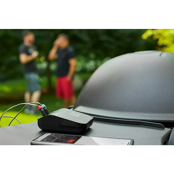 Weber Smart-Grill-Assistent - L x B x H: 9,8 x 3,4 x 7,5 cm - Smartfire-Grillthermometer auf einem Grill mit zwei Personen im Hintergrund. Das Gerät verfügt über farbcodierte Sensoren und ein Smartphone.