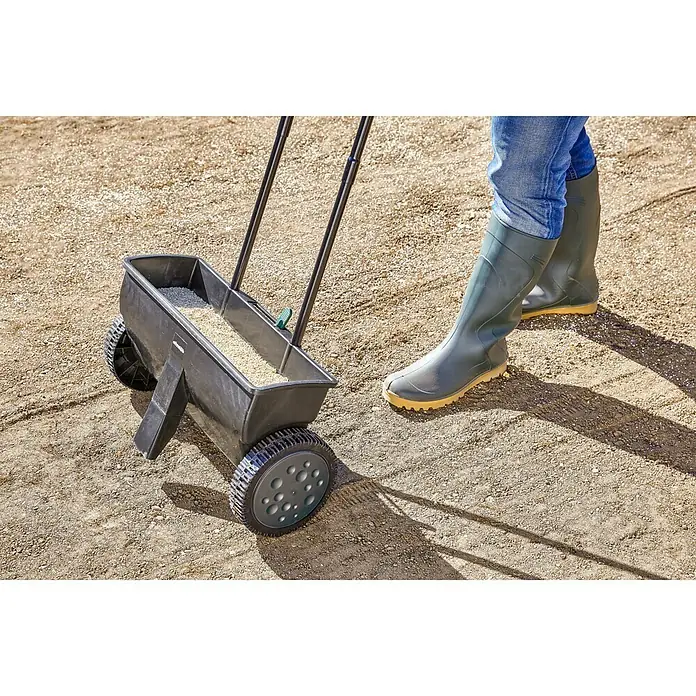 Gardol Carro esparcidor ST 451 - 12 l, Plástico - Imagen de un esparcidor de semillas de césped en uso, con una persona en botas de agua empujándolo.