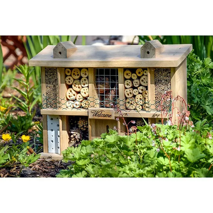Dobar Insektenhotel Landsitz Superior - 47 x 12,5 x 34 cm, Eiche - Ein charmantes Insektenhotel aus Holz, eingebettet in üppiges Grün, mit kleinen Holzdetails und einer einladenden "Welcome"-Aufschrift.
