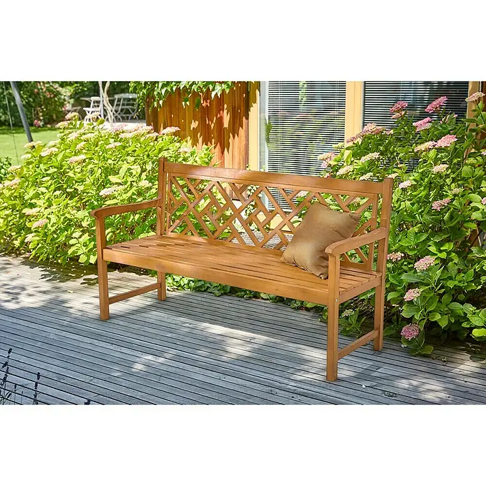 Sunfun Gartenbank - B x T x H: 158 x 61,5 x 91 cm, Akazie, Naturbraun - Eine hölzerne Gartenbank mit einem dekorativen Rückendesign und einem Kissen, die auf einer Holzterrasse steht und von Pflanzen umgeben ist.