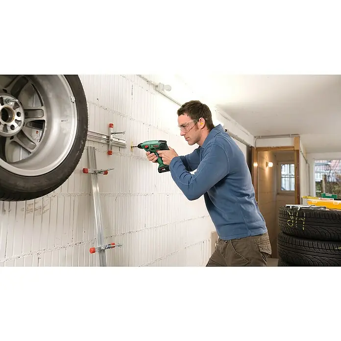 Bosch Power for All 18V Akku-Bohrhammer Uneo Maxx - 18 V, 1 Akku, 0,6 J - Mann montiert eine Reifenhalterung mit einem grünen Akkuschrauber an einer Wand in einer Garage.