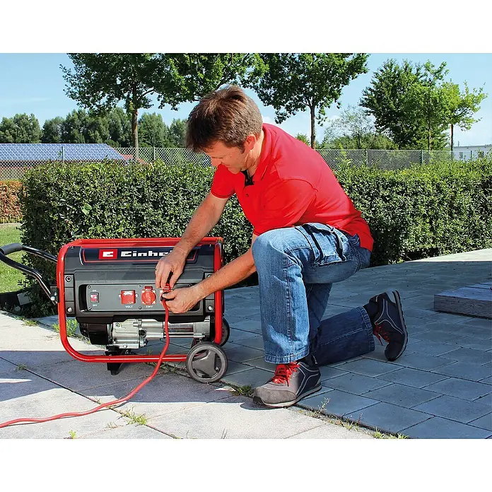 Einhell Stromerzeuger TC-PG 35/E5 - Tankvolumen: 15 l - Ein Mann schließt einen roten Einhell Stromerzeuger an. Der Generator steht auf Steinplatten vor einer grünen Hecke im Freien.