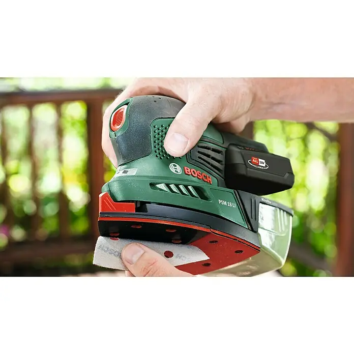 Bosch Power for All 18V Lijadora delta de batería PSM 18 Li - 18 V, Iones de litio, Sin batería, 22.000 r.p.m. - Primer plano de una lijadora multifunción inalámbrica Bosch PSM 18 LI con papel de lija adjunto, sostenida por una mano, sobre un fondo borroso.