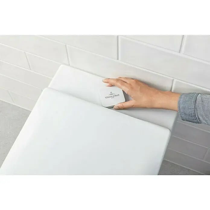 Villeroy & Boch Subway 2.0 Wand-WC ViFresh - Spülrandlos, Ohne Beschichtung, Spülform: Tief, WC Abgang: Waagerecht, Weiß - Eine Hand legt einen Villeroy & Boch WC-Duftstein auf einen weißen Toilettenspülkasten, um für Frische im Badezimmer zu sorgen.
