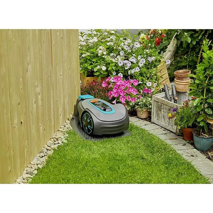 Gardena Mähroboter Sileno minimo 500 - 500 m², Schnittbreite: 16 cm, Induktionsgestützte Navigation, Begrenzungskabel - Ein Gardena Mähroboter parkt in einer grünen Gartenecke, umgeben von Blumen und einem Holzzaun. Das Gerät ist grau mit blauen Akzenten.