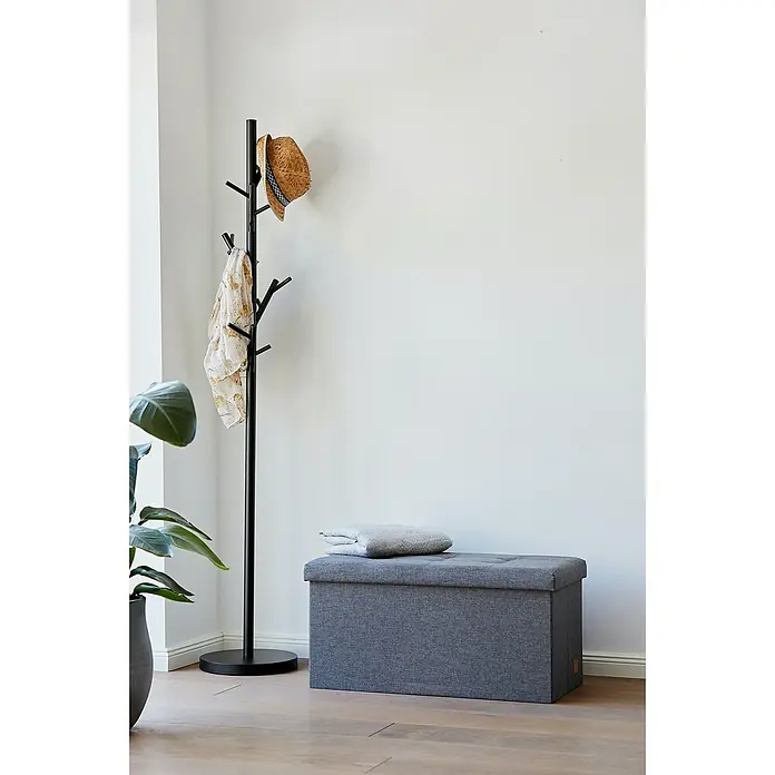 Store It Premium Sitz- & Aufbewahrungsbox Home - L x B x H: 38 x 76 x 38 cm, Grau - Grauer, gepolsterter Hocker mit Deckel und ein Garderobenständer aus Metall mit Hut und Schal stehen neben einer Zimmerpflanze im Raum.