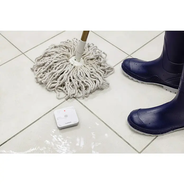 Bosch Smart Home Funk-Wassermelder - Batteriebetrieben, 75 x 75 x 25 mm - Das Bild zeigt einen Bosch Wassermelder, der auf einem gefliesten Boden neben einem Mopp und Gummistiefeln liegt. Der Sensor überwacht Wasseraustritte.