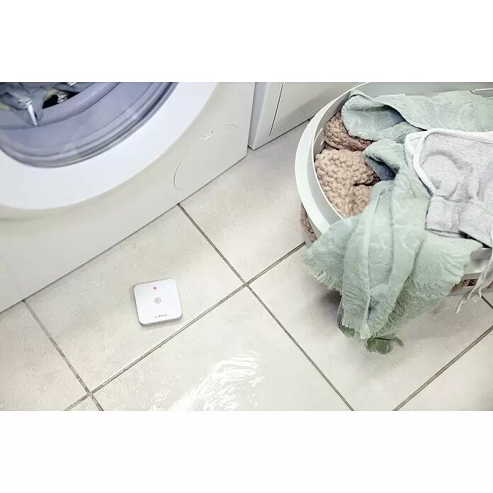 Bosch Smart Home Funk-Wassermelder - Batteriebetrieben, 75 x 75 x 25 mm - Weißer Bosch Wasserlecksensor auf Fliesenboden, entdeckt kleine Pfütze nahe Waschmaschine.