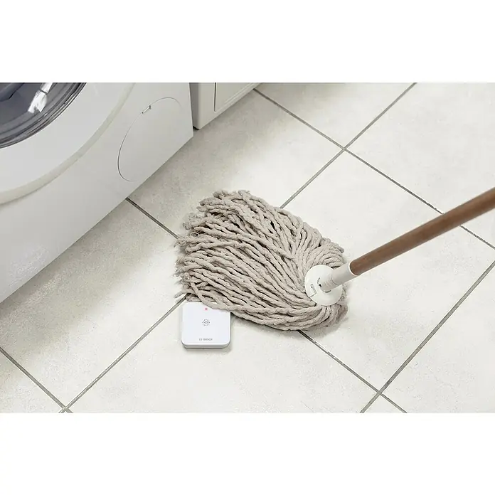 Bosch Smart Home Funk-Wassermelder - Batteriebetrieben, 75 x 75 x 25 mm - Ein Mopp und ein Bosch Smart Home Bewegungsmelder auf einem hellen Fliesenboden neben einer Waschmaschine.