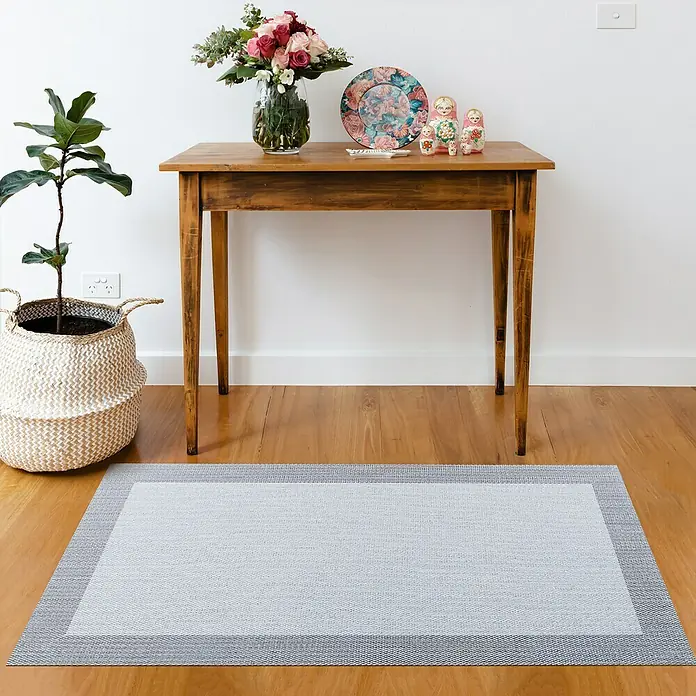 Alfombra de vinilo Living - Gris, 150 x 80 cm - Una alfombra gris con un borde de dos tonos se coloca debajo de una mesa de madera con flores y muñecas matrioska en una habitación luminosa.