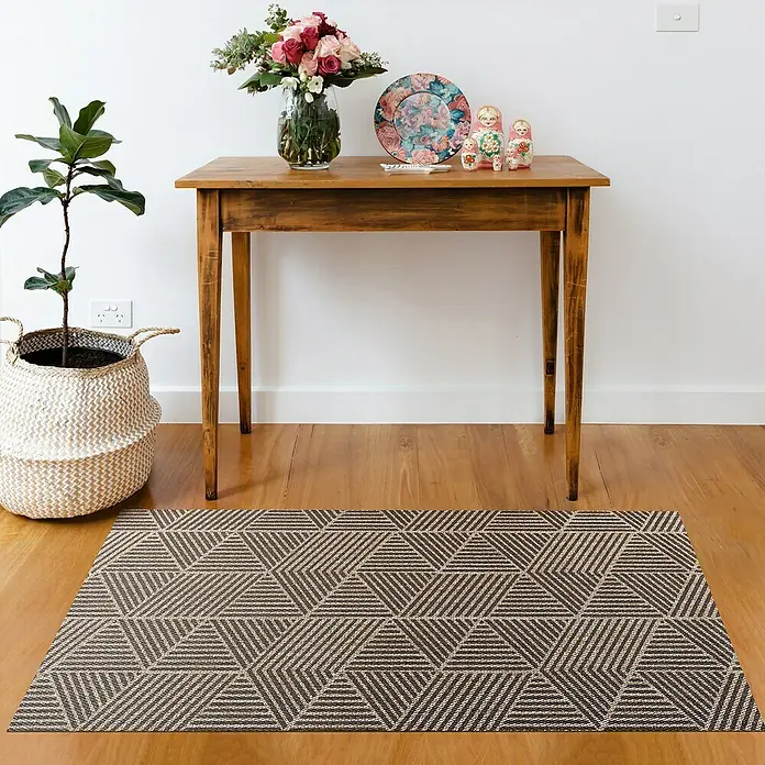 Alfombra de vinilo Living Geométrica - Beige/Marrón, 290 x 200 cm - Una alfombra con un patrón geométrico en tonos marrones y beige está colocada debajo de una mesa de madera, junto a una planta en maceta y varias decoraciones.
