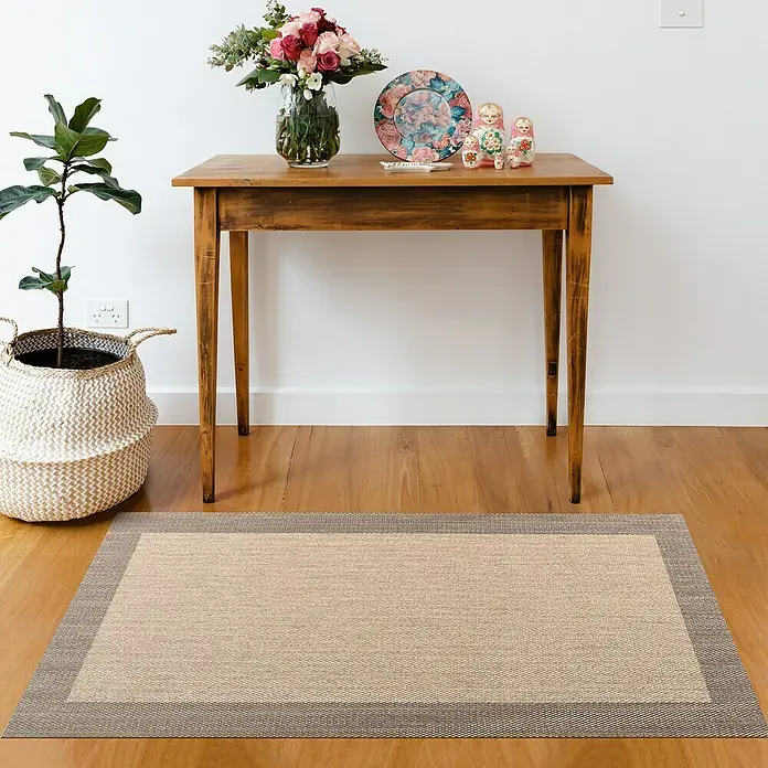 Alfombra de vinilo Living - Beige/Tostado, 200 x 60 cm - Una alfombra beige con borde marrón está colocada sobre un piso de madera frente a una mesa de madera con flores, un plato y muñecas sobre ella.