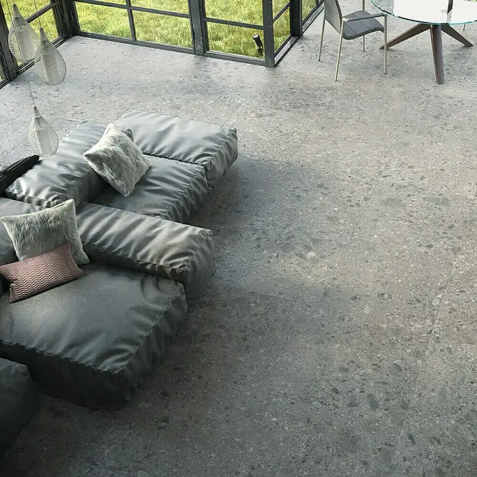 Feinsteinzeugfliese Hannover Steel - 60 x 60 cm, Grau, Matt - Graue Terrazzo-Fliesen bedecken den Boden eines modernen Wohnraums mit Fenster, Tisch und einer modularen Couch.