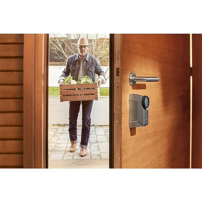 Abus Set de cerradura electrónica HomeTec Pro - 2 pzs. - Un hombre con un sombrero y una caja de verduras se encuentra frente a una puerta con una cerradura inteligente y manija.