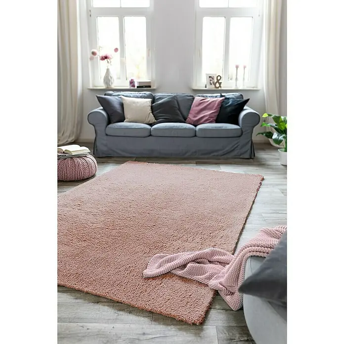 Hochflorteppich Sugar - Rosa, 180 x 120 cm, 100 % Polyester - Ein weicher, rosafarbener Teppich liegt in einem hellen Wohnzimmer mit einem grauen Sofa, Kissen und einem Strickpouf.
