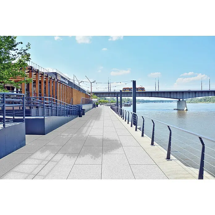 Terrassenfliese Cera 3.0 - 40 x 40 x 3 cm, Granito Montorfano, Matt - Aufnahme einer Uferpromenade mit einem Weg, der mit quadratischen grauen Steinen gepflastert ist, entlang eines Flusses unter einer Brücke.