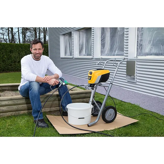 Wagner High Efficiency Airless Sistema de pulverización de pintura Control Pro 350 M Set - Consumo de potencia: 600 W, Caudal: 1,5 l/min - Un hombre está sentado junto a un pulverizador de pintura Wagner en un patio de madera. El pulverizador está sentado sobre cartón.