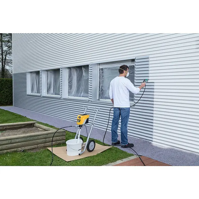 Wagner High Efficiency Airless Farbsprühsystem Control Pro 350 M Set - Leistungsaufnahme: 600 W, Förderleistung: 1,5 l/min - Ein Mann streicht eine Hauswand mit einer Wagner Farbspritzpistole, während Fenster abgeklebt sind. Der Farbbehälter steht auf dem Rasen.