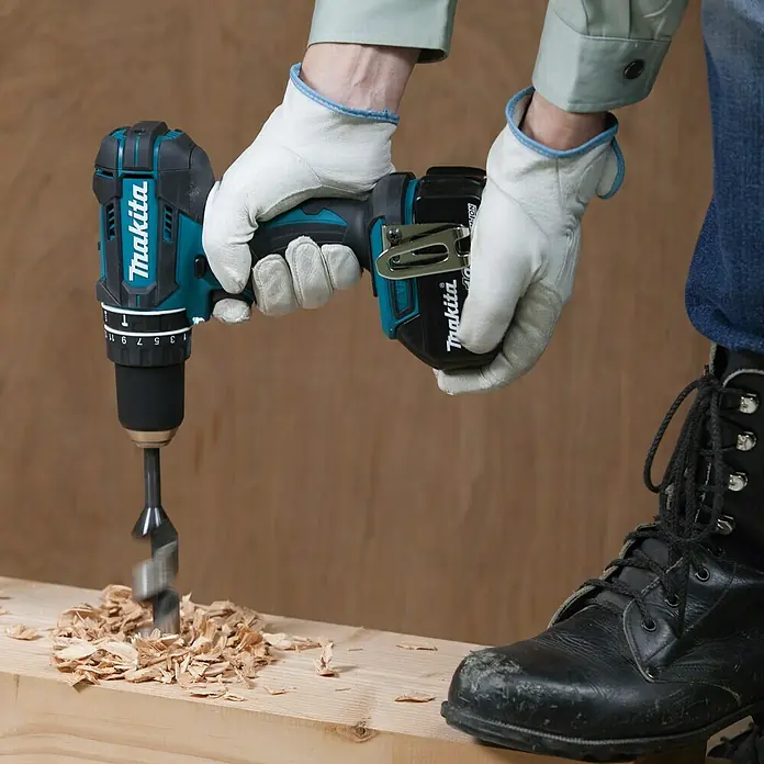Makita Set taladro percutor de batería DTD152Z + taladro combinado DHP482Z - 18 V, 3 baterías, 3 Ah - Primer plano de un taladro inalámbrico Makita perforando madera, con virutas de madera y un par de guantes.