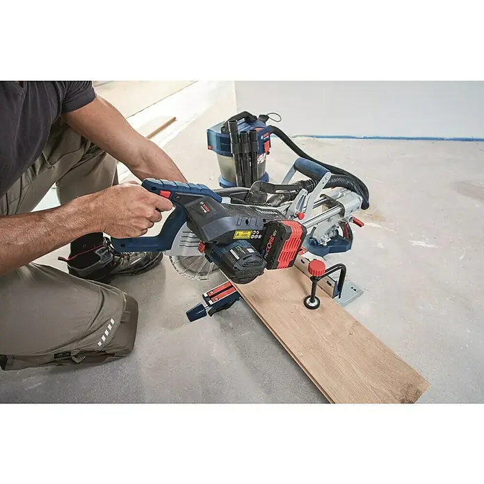 Bosch Professional AMPShare 18V Akku-Kapp- & Gehrungssäge 18V-216 Solo - 18 V, Ohne Akku, Durchmesser Sägeblatt: 216 mm - Eine Bosch Akku-Kapp- und Gehrungssäge mit rotem Akku schneidet ein Stück Holz, während eine Person sie bedient und ein Staubsauger daneben steht.