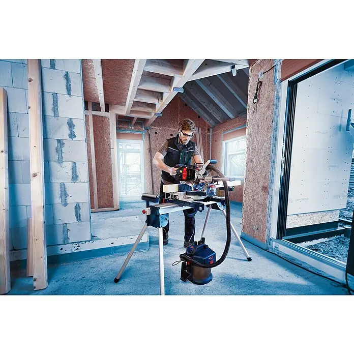 Bosch Professional AMPShare 18V Akku-Kapp- & Gehrungssäge 18V-216 Solo - 18 V, Ohne Akku, Durchmesser Sägeblatt: 216 mm - Ein Mann bedient eine Bosch Kapp- und Gehrungssäge auf einer Baustelle, angeschlossen an einen Staubsauger, um Holz präzise zu schneiden.