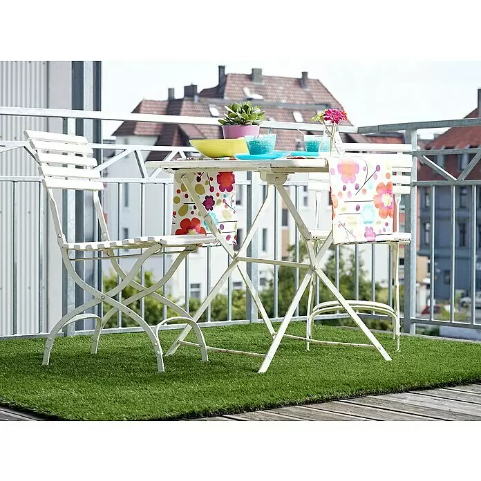Kunstrasen Meterware Koh Samui - Breite: 400 cm, Grün, Mit Drainagenoppen - Weißer klappbarer Balkontisch und zwei Stühle auf Kunstrasen, dekoriert mit farbigen Schüsseln und einer Pflanze, bereit für den Außenbereich.