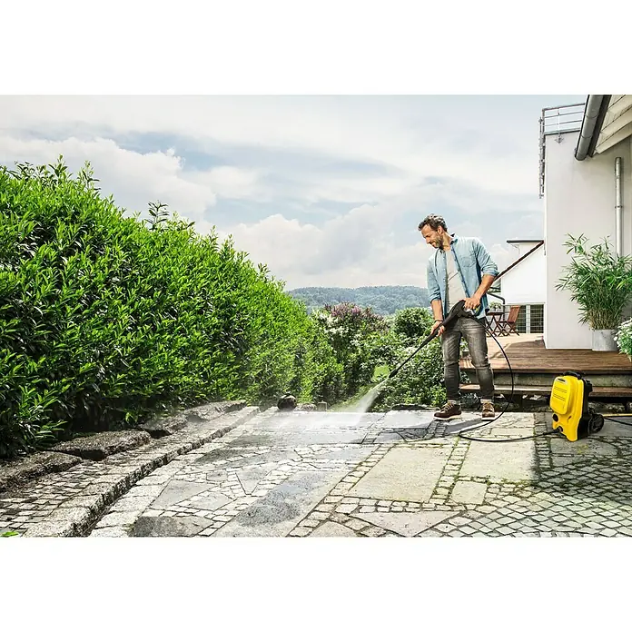 Kärcher Hidrolimpiadora de alta presión K4 Compact UM - 130 bar, 1.800 W, Capacidad de bombeo: 420 l/h - Un hombre limpia un patio de piedra con una hidrolimpiadora Kärcher amarilla, creando un camino limpio.