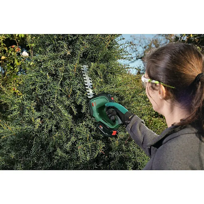 Bosch Power for All 18V Akku-Gras- & Strauchschere AdvancedShear 18V-10 - 18 V, Li-Ionen, 1 Akku, Schwertbreite: 10 cm - Eine Frau schneidet eine Hecke mit einer grünen Akku-Heckenschere von Bosch. Die Heckenschere ist gut sichtbar und wird aktiv benutzt.